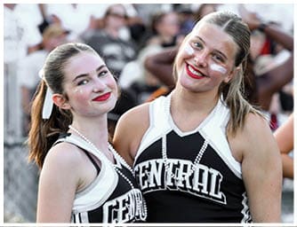 Two adults wearing cheerleading uniforms with “CENTRAL" on the front smile with face paint and beads while standing outdoors among a crowd. Descripci n generada con IA