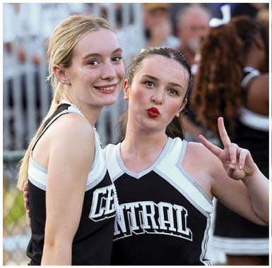 Two people wearing cheerleading uniforms are standing close together outdoors with one smiling and the other making a playful expression. Descripci n generada con IA