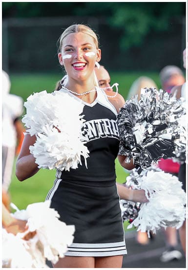 A person wearing a cheerleading uniform with face paint is smiling and holding white and silver pom-poms outdoors. Descripci n generada con IA