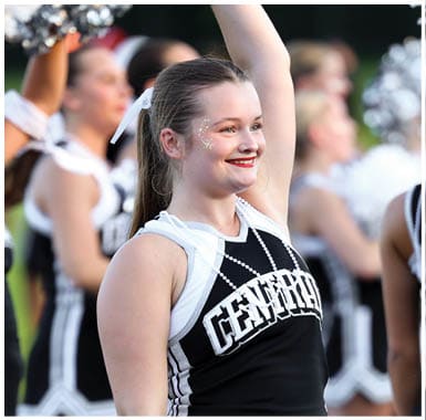 A person in a cheerleading uniform with a ponytail and face glitter is smiling and raising a pom-pom while other cheerleaders stand in the background. Descripci n generada con IA