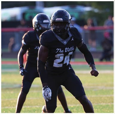 Two people in football uniforms are actively engaged on a field with one person in red preparing to block and two people in black moving defensively. Descripci n generada con IA