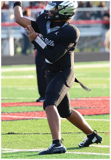 A person wearing a black football uniform and helmet is preparing to throw a football on a green field with spectators in the background. Descripci n generada con IA