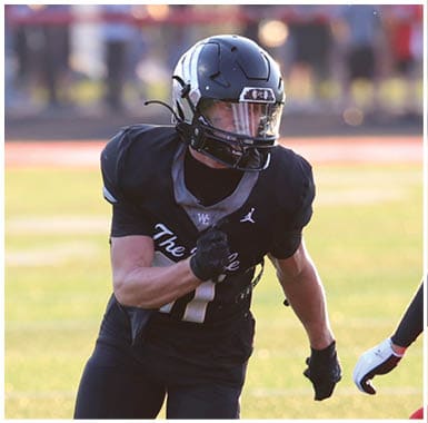A person in black sports gear is running with a football while two people in red sports uniforms prepare to block or tackle on a sunlit football field. Descripci n generada con IA