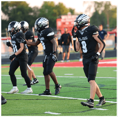 People in black football uniforms and helmets stand on a green field with white yard lines and a red end zone in the background. Descripci n generada con IA