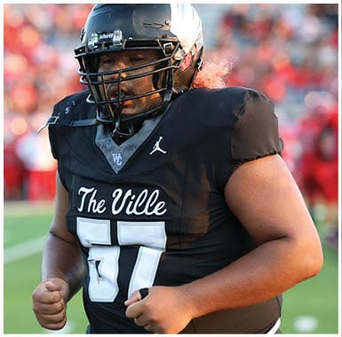 A person wearing a black football uniform with “The Ville" and the number 57 on the jersey is standing on a field with a helmet and faceguard. Descripci n generada con IA