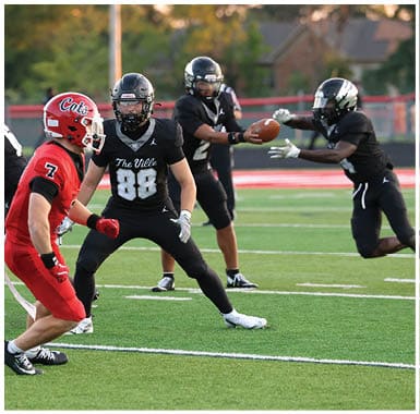 People in football uniforms are actively engaged in a game on a green field with one player in red running while others in black attempt to block or tackle. Descripci n generada con IA
