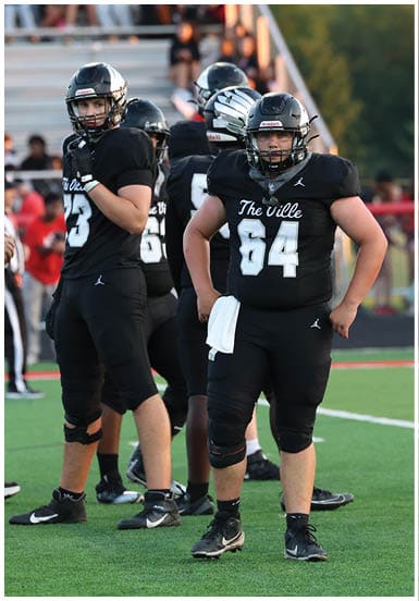 Two people in black football uniforms with helmets stand on a green field during a game or practice, with blurred spectators and other players in the background. Descripci n generada con IA