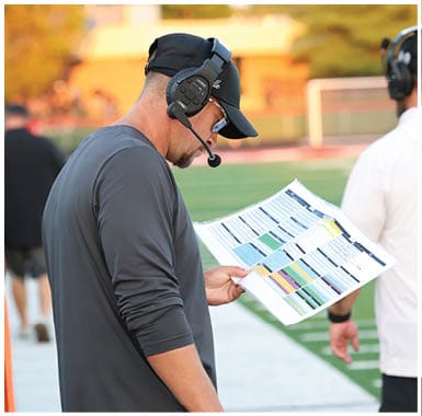 A person wearing a headset and dark long-sleeve shirt is standing on a sports field looking at a laminated play sheet while others stand nearby. Descripci n generada con IA