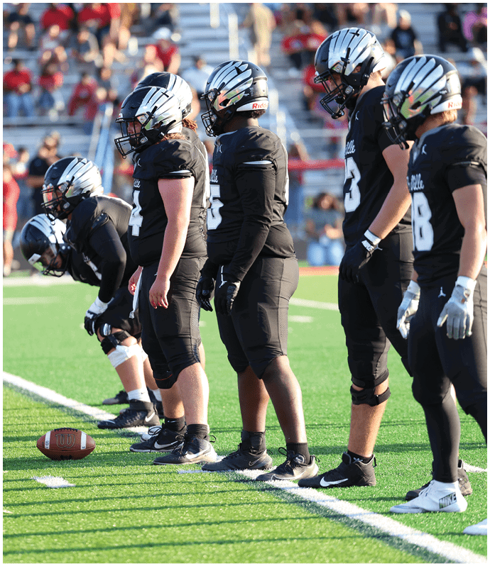 Five people in black football uniforms and silver helmets stand in a line on a green field preparing for a play. Descripci n generada con IA