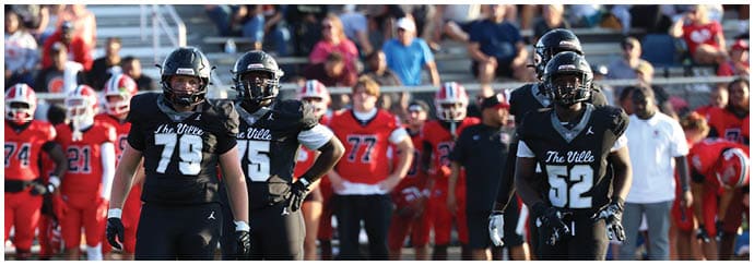 People in football uniforms stand on a field with a crowd of spectators in the background. Descripci n generada con IA