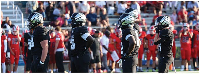 People in black sports uniforms stand on a field facing a group of people in red uniforms with a crowd seated in bleachers behind them. Descripci n generada con IA