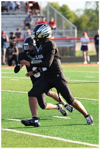 Two people in black football uniforms and helmets are actively playing on a green field with spectators in the background. Descripci n generada con IA