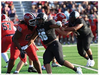 Two people in football uniforms are engaged in a play on a green field with a crowd in the background. Descripci n generada con IA