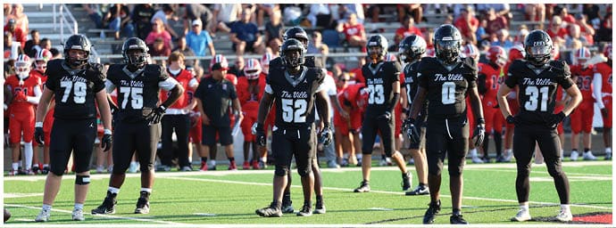 A group of people wearing black football uniforms with helmets stand on a field facing the camera with a crowd of spectators in the background. Descripci n generada con IA