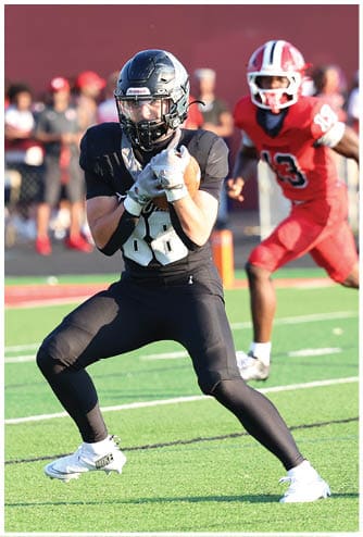 A person in black sports gear catches a football on a green field while another person in red sports gear runs behind them. Descripci n generada con IA