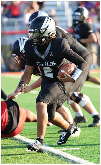 A person wearing a black football uniform with the number 2 is running with a football on a green field while other players in black and red uniforms are around them. Descripci n generada con IA
