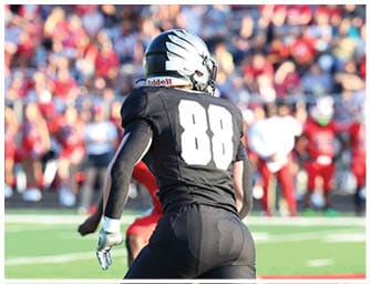 Two people in football uniforms are running on a field with one wearing red and the other wearing black. Descripci n generada con IA