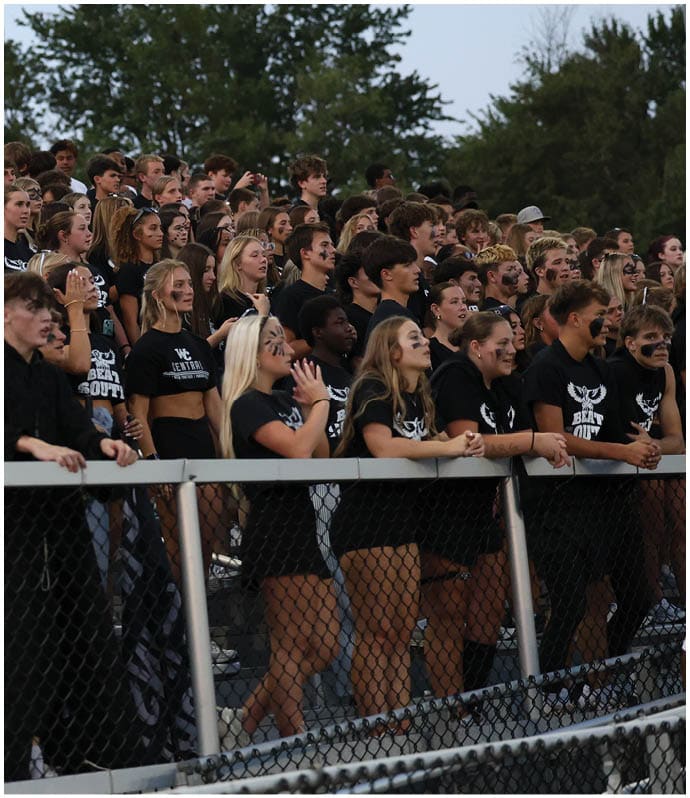 A large group of people dressed mostly in black with face paint are standing closely together in bleachers, some cheering and one holding a large orange cone. Descripci n generada con IA