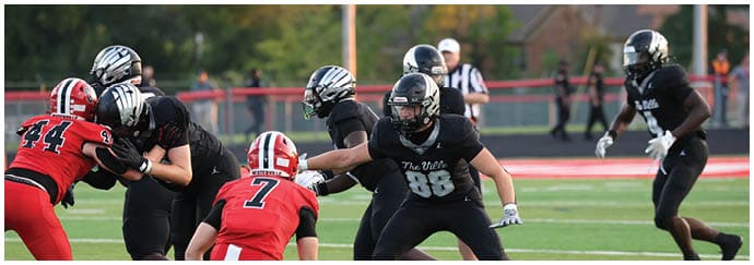 Adults in football uniforms are engaged in a game on a field with some players in black and others in red preparing for a play. Descripci n generada con IA