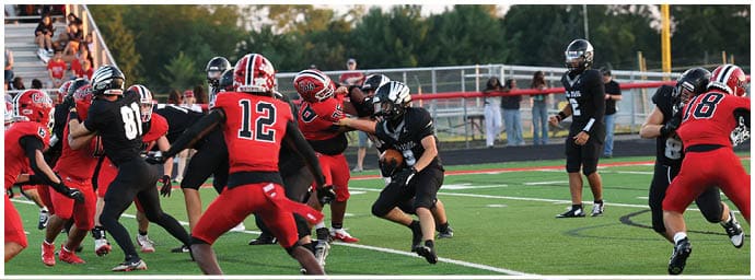 People wearing red and black football uniforms are actively playing on a green field with one person running with the ball while others are preparing to block or tackle. Descripci n generada con IA