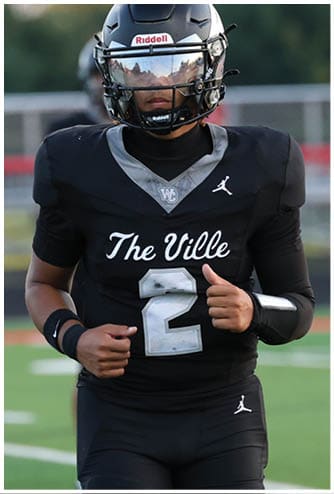A person in a black football uniform with the number 2 and “The Ville" on the jersey is jogging on a football field with two other players in the background. Descripci n generada con IA