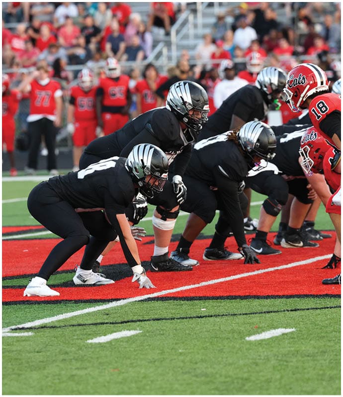 Several people in black sports uniforms and helmets are lined up in a crouched position on a football field facing opponents in red uniforms during a game. Descripci n generada con IA