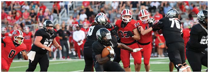 Players in black and red football uniforms are actively engaged in a game on a green field with a crowd in the background. Descripci n generada con IA