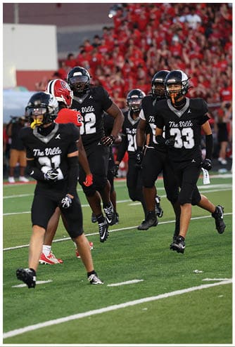 People in black football uniforms with “The Ville" on the front are running on a football field with a crowd in the background. Descripci n generada con IA