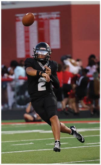 A person in a black sports uniform is throwing a football on a green field while another person in a red sports uniform approaches. Descripci n generada con IA