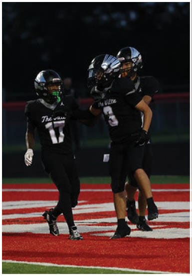 Three people wearing black football uniforms and helmets are engaged in a game or practice on a field at dusk or night. Descripci n generada con IA