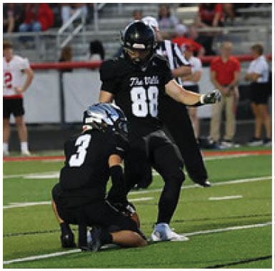 Two people in black football uniforms are on a field with one holding the ball for the other to kick during a game with spectators in the background. Descripci n generada con IA