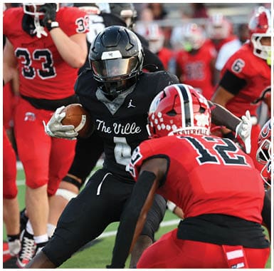 A group of people in red and black football uniforms are actively playing on a green field with one person in black holding the ball while others in red attempt to block or tackle. Descripci n generada con IA