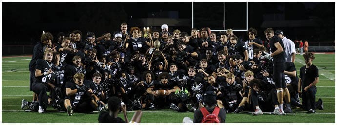 A large group of people wearing matching dark sports uniforms are gathered closely together on a field at night, celebrating and holding up a trophy. Descripci n generada con IA
