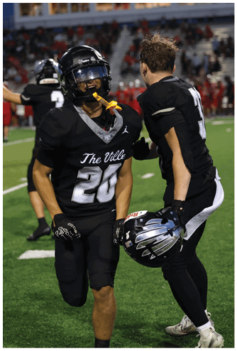 Two people in black football uniforms are on a green field, one holding a helmet and the other with a mouthguard hanging from their face. Descripci n generada con IA