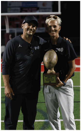 A person with short light hair wearing a black polo shirt is smiling and holding a large football-shaped trophy on a sports field at night. Descripci n generada con IA