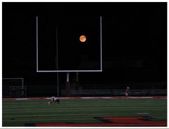 A bright orange full moon is visible in a dark night sky next to a tall, thin pole. Descripci n generada con IA
