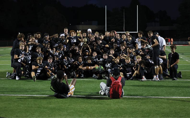 A large group of people wearing matching dark sports uniforms pose together on a football field at night while two people sit on the grass facing them. Descripci n generada con IA