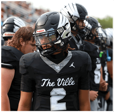 A person wearing a black and gray football uniform with a helmet and visor is on a field with another similarly dressed person in the background. Descripci n generada con IA