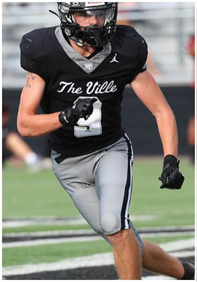 An adult wearing a black football jersey with “The Ville" written on it and gray pants is running on a football field. Descripci n generada con IA