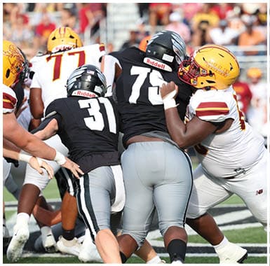 Two people in black sports uniforms are being blocked by a person in a white and yellow football uniform on a green field with a crowd in the background. Descripci n generada con IA