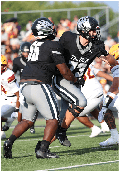 Two people in black and gray football uniforms are engaged in a play on a grassy field with other players in the background. Descripci n generada con IA
