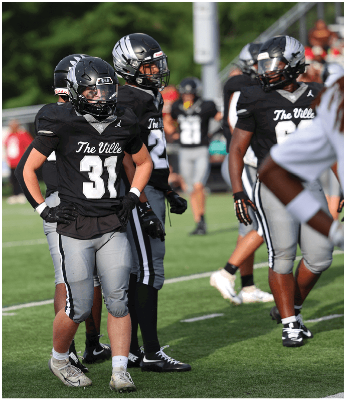 Several people in black and gray football uniforms with helmets stand on a grassy field during a game or practice. Descripci n generada con IA