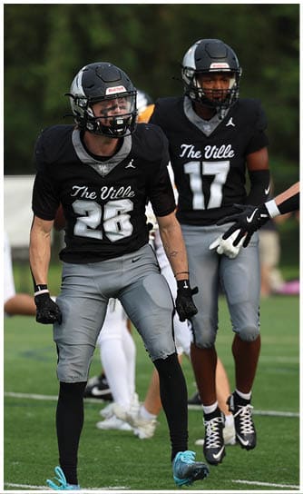 A person wearing a black football jersey with the number 26 and gray pants is standing on a football field with other players in the background. Descripci n generada con IA