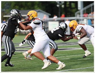 Two people in white football uniforms with yellow helmets are engaged in a play against a person in a black football uniform with a black and white helmet on a green field. Descripci n generada con IA