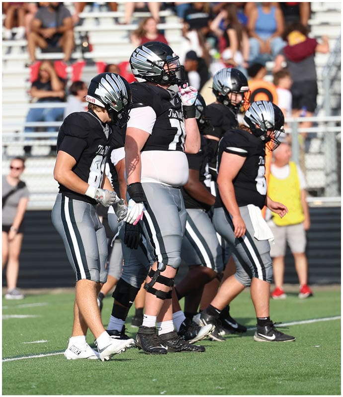 Several people wearing black and gray football uniforms stand on a green field with two people in casual clothing visible in the background near bleachers. Descripci n generada con IA