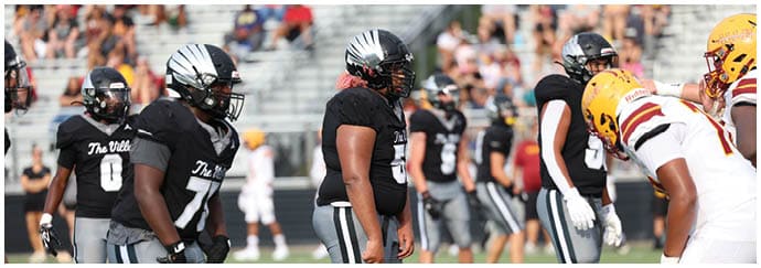 People in football uniforms from two opposing teams are lined up facing each other on a field with spectators in the background. Descripci n generada con IA
