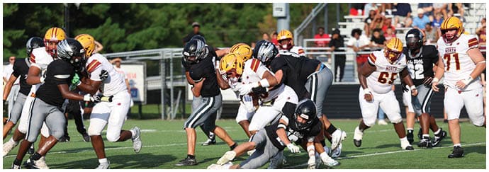 People wearing football uniforms are actively engaged in a game on a grassy field with some players tackling and others running. Descripci n generada con IA