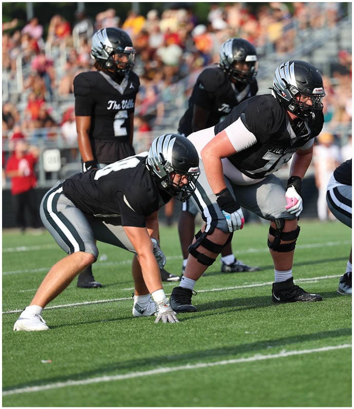 Several people in football uniforms and helmets are positioned on a grassy field, preparing for a play during a game. Descripci n generada con IA