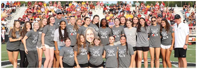 A group of people wearing matching gray t-shirts and athletic shorts pose together on a sports field with a crowd in the background. Descripci n generada con IA