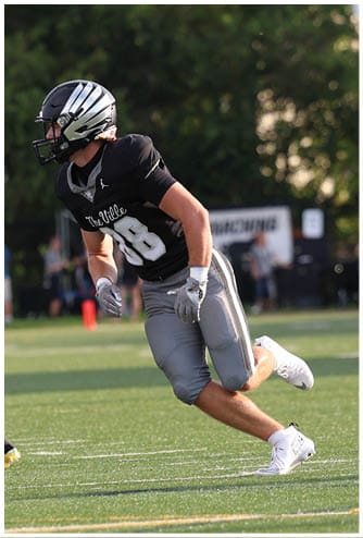 A person wearing a black helmet, black shirt, gray pants, and white shoes is running on a green football field with other people and trees in the background. Descripci n generada con IA
